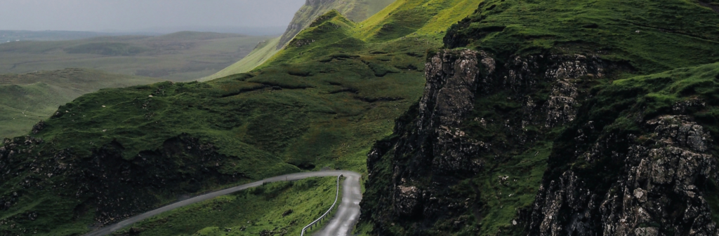 A winding road curves through green hills and rocky cliffs under a cloudy, overcast sky.