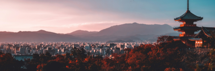 A cityscape with a traditional Japanese pagoda in the foreground and mountains in the background during sunset, illustrating a serene blend of cultural heritage and sustainable business travel opportunities amidst stunning natural scenery. | Dyme