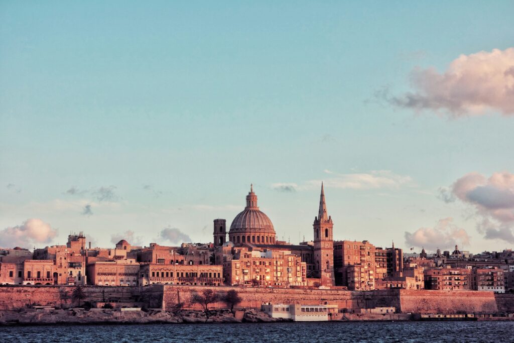 City skyline with historic buildings and a large dome by the water under a partly cloudy sky at sunset.