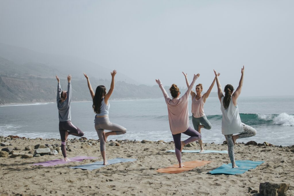 Five people practice yoga in tree pose on mats on a sandy beach by the ocean, with mountains in the background.