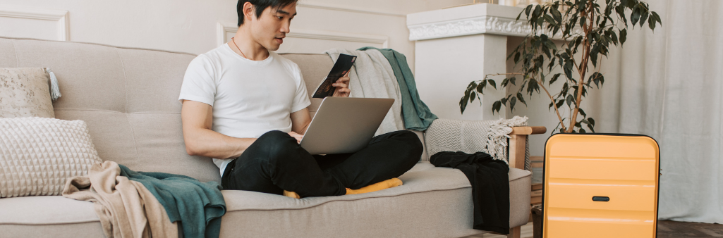 Man sitting on a couch with a laptop, looking at his phone, yellow suitcase nearby.