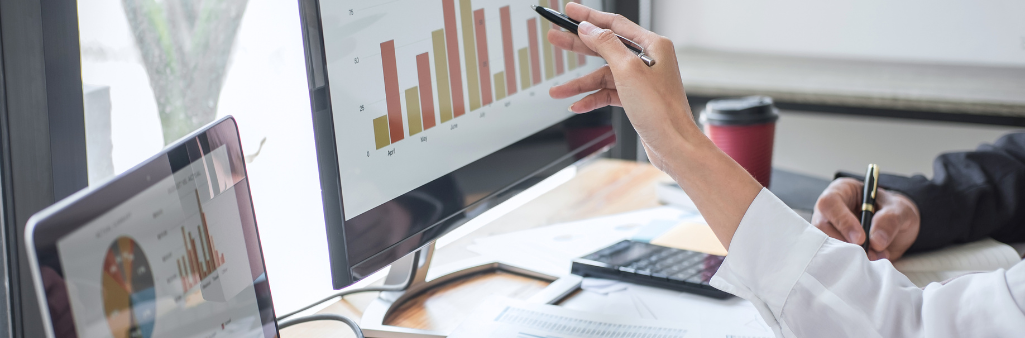 Two people analyzing bar graphs on computer screens at a desk with documents, a calculator, and a coffee cup.