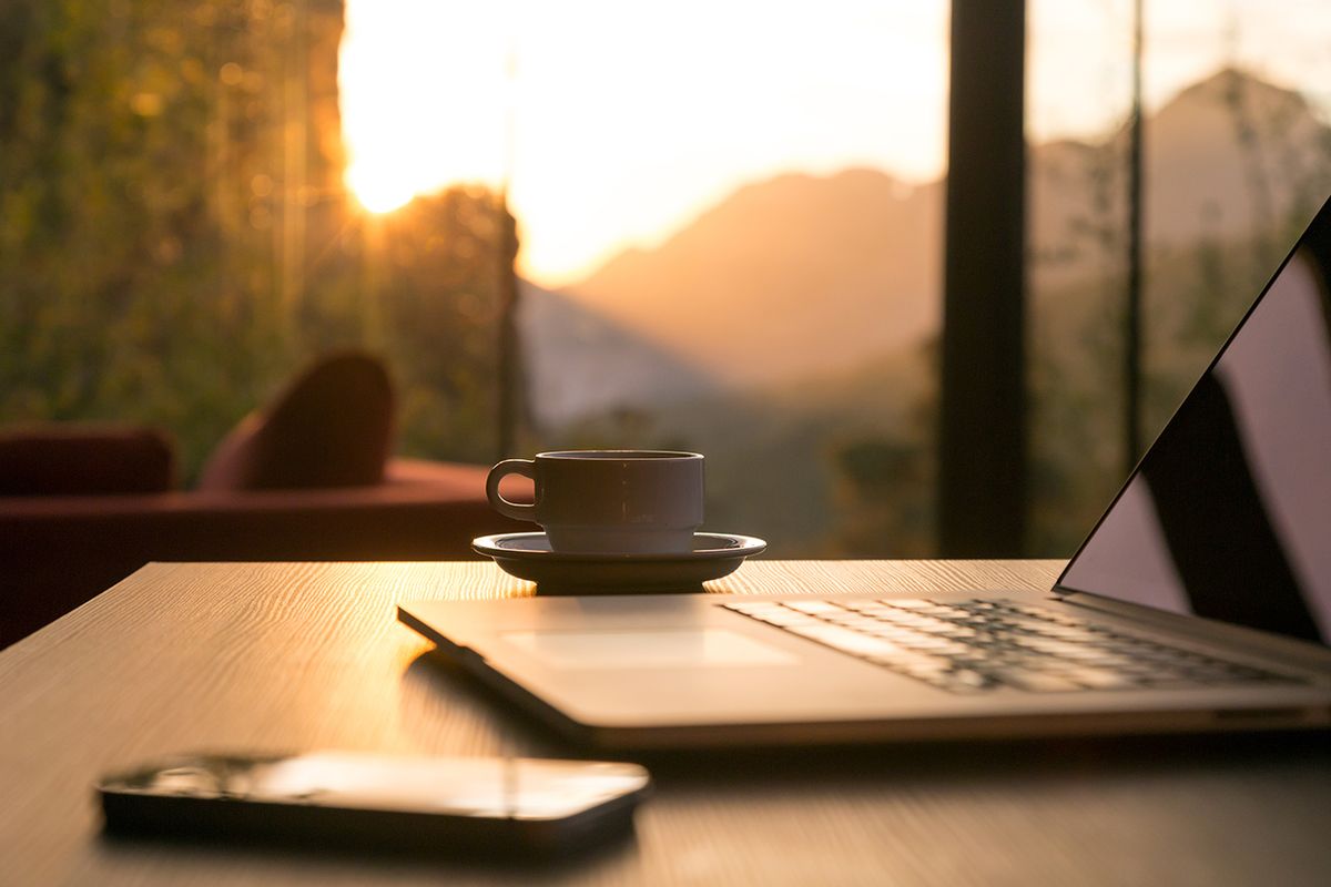 Coffee cup and open laptop on a wooden desk by a window with a mountain sunset, evoking remote business travel