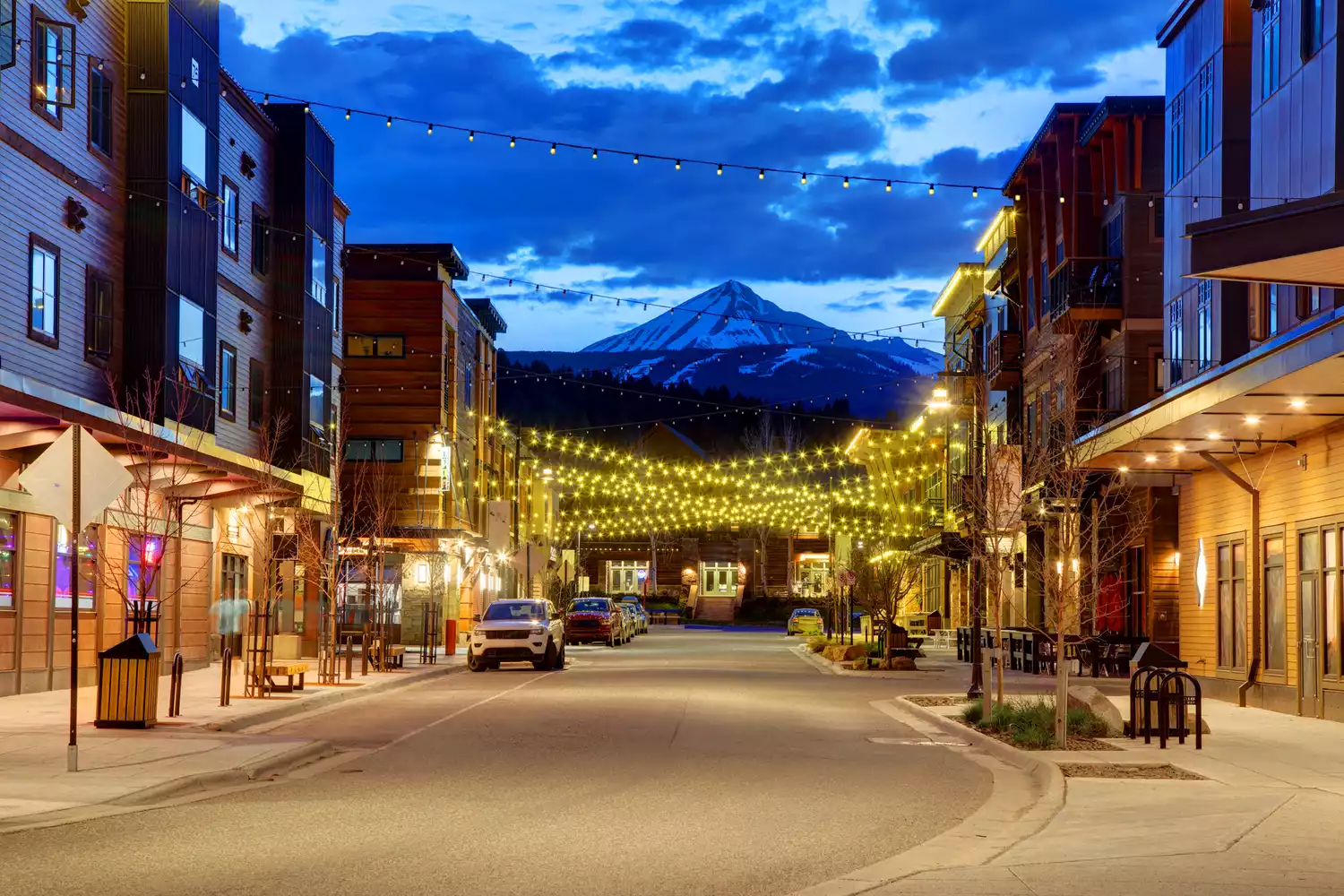 Mountain resort town street at dusk with string lights overhead and a snow-capped peak in the distance