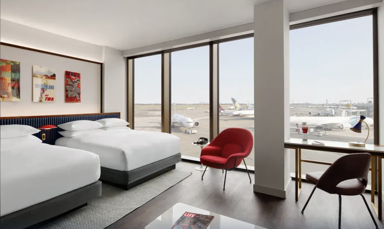 TWA Hotel guest room at JFK with twin beds, red Womb chairs and floor-to-ceiling windows over the runway
