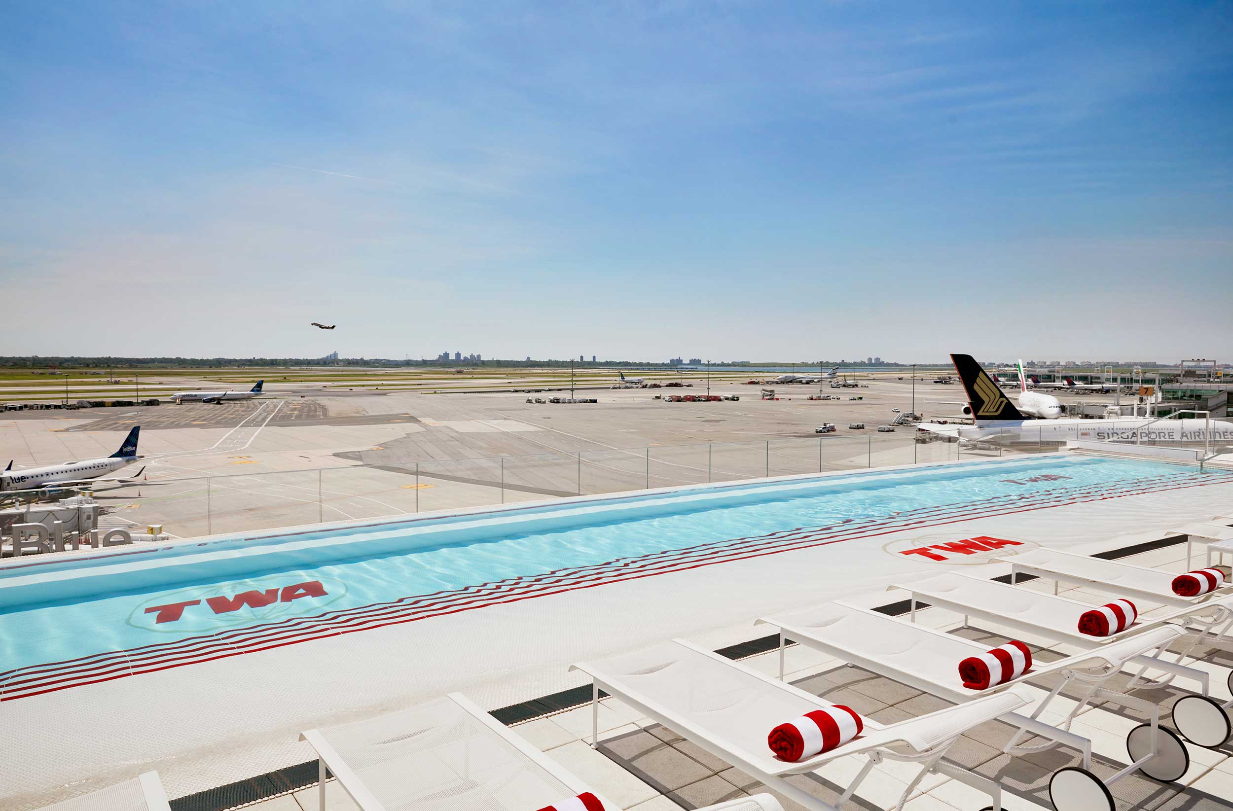 TWA Hotel rooftop pool at JFK with red-striped loungers overlooking the airfield and taxiing aircraft