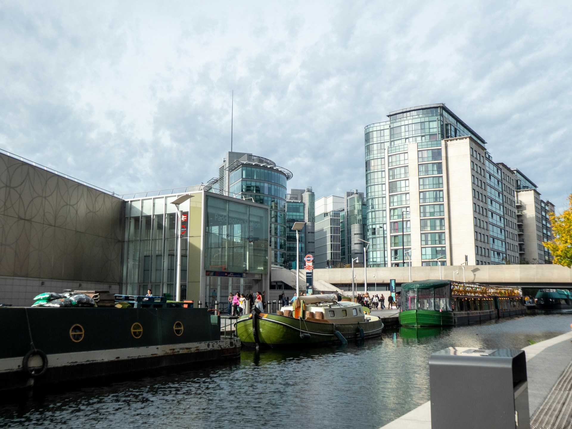 Paddington Basin waterway in London with moored canal barges and glass office and apartment towers under a cloudy sky
