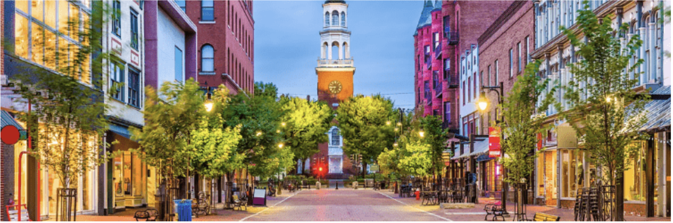 Tree-lined downtown pedestrian street at dusk with a white church tower in the distance