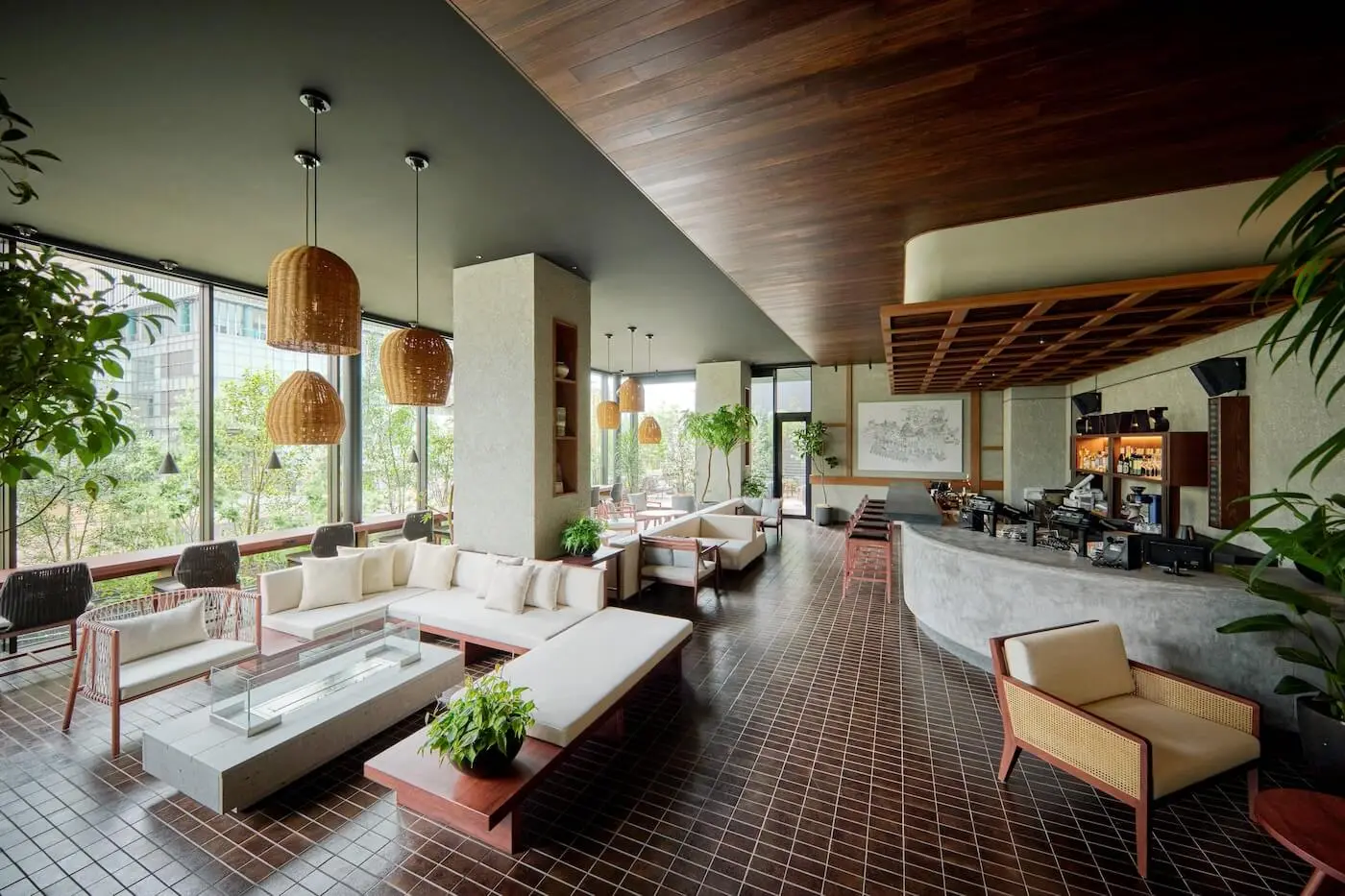 Lounge of a Fukuoka hotel with a sloped wood ceiling, rattan pendant lights, cream sofas, a curved bar and large windows to greenery
