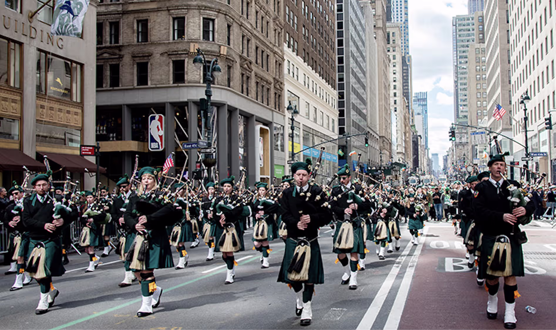 Bagpipers in green kilts marching up Fifth Avenue past Manhattan buildings during the NYC St. Patrick's Day Parade