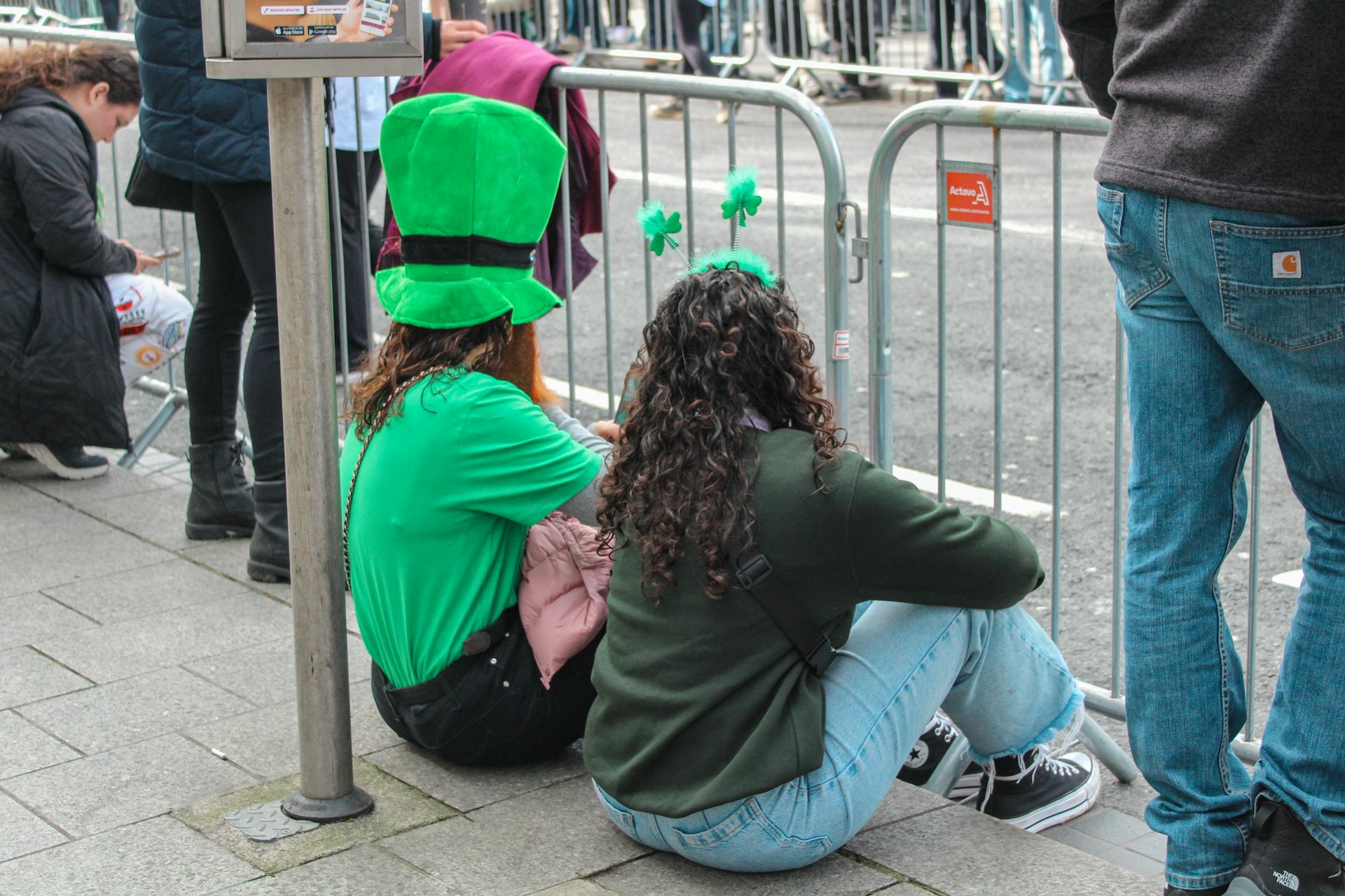 Two people in green St. Patrick's Day hats sitting by a crowd-control barrier along a parade route in Chicago