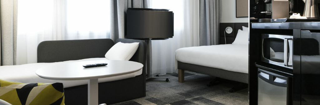 Modern hotel room with a bed, sofa, round table, TV, microwave, and mini fridge near large windows.