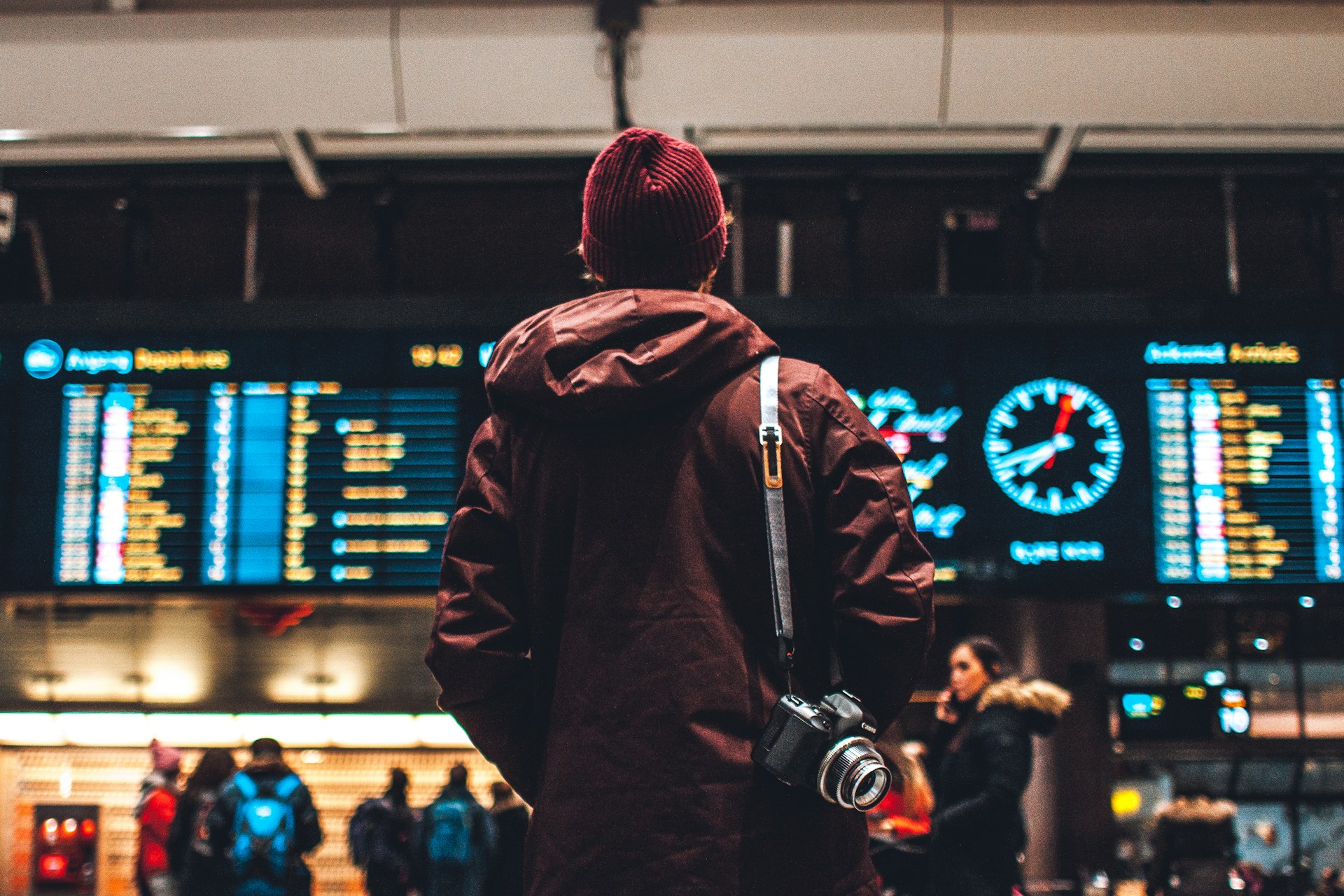 Traveler in a maroon parka with a camera facing a lit departures board and clock in a transit hub while planning a multi-city March Madness route