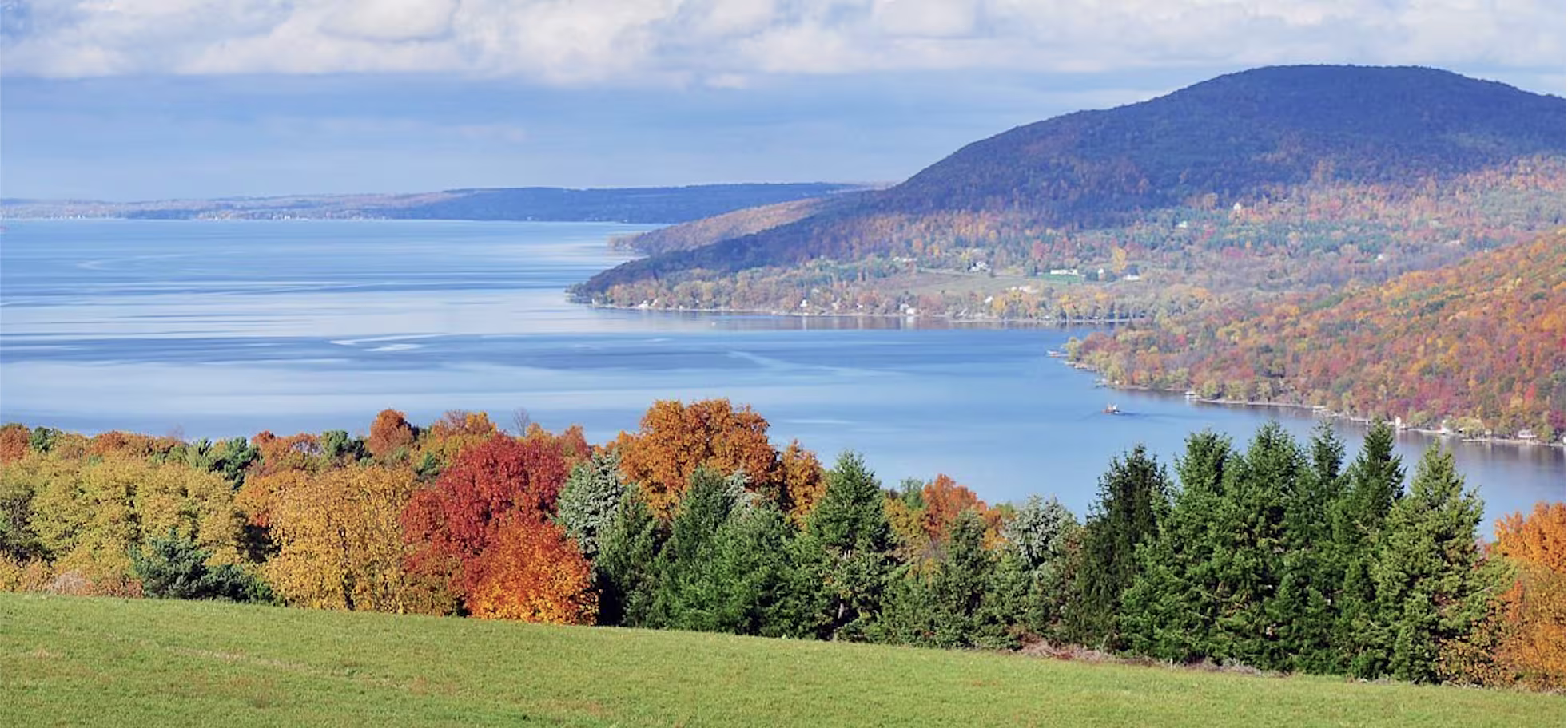 Lake winding between forested hills with autumn foliage and a green meadow in the foreground, a quiet Memorial Day getaway scene