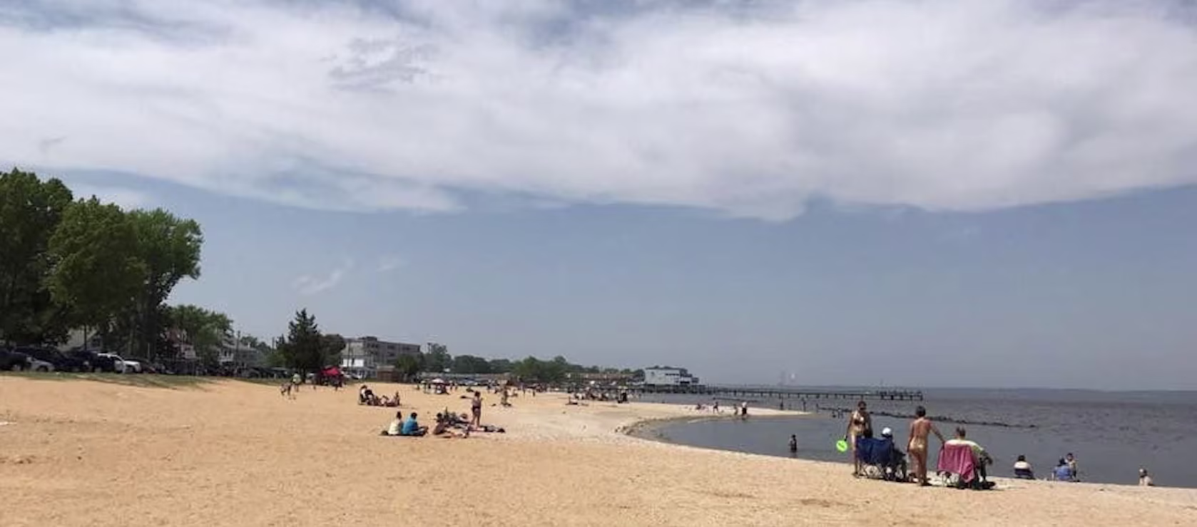 Sandy bay beach with scattered visitors, beach chairs and a pier under a partly cloudy sky on a warm holiday weekend