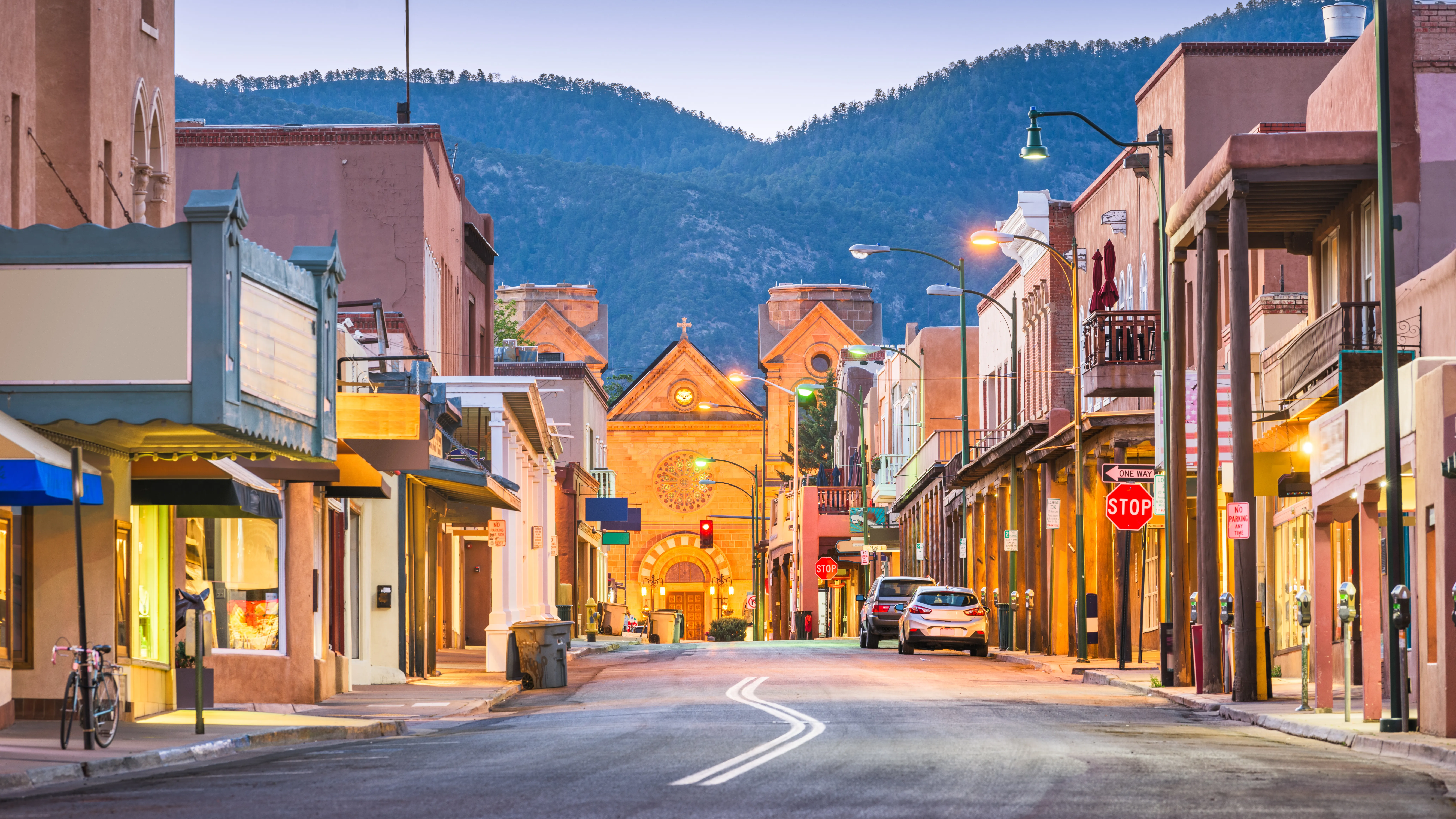 Twilight street in Santa Fe, New Mexico lined with adobe shops, parked cars and an old mission church framed by mountains