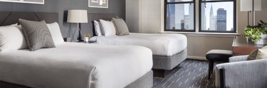 Modern hotel room with two double beds, large window, city view, desk, lamp, and neutral decor.
