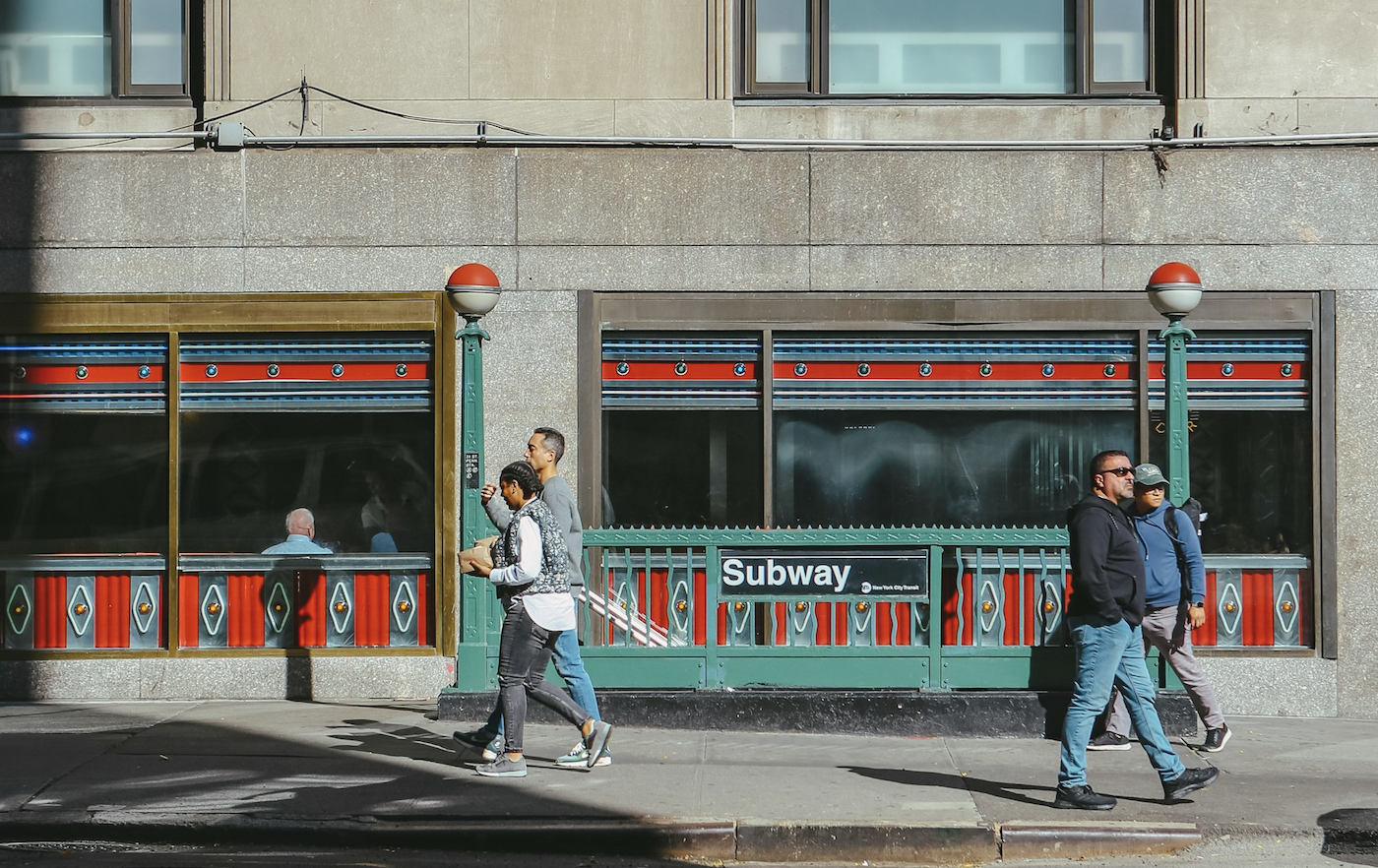 Pedestrians walking past a New York City subway station entrance on a sunny day