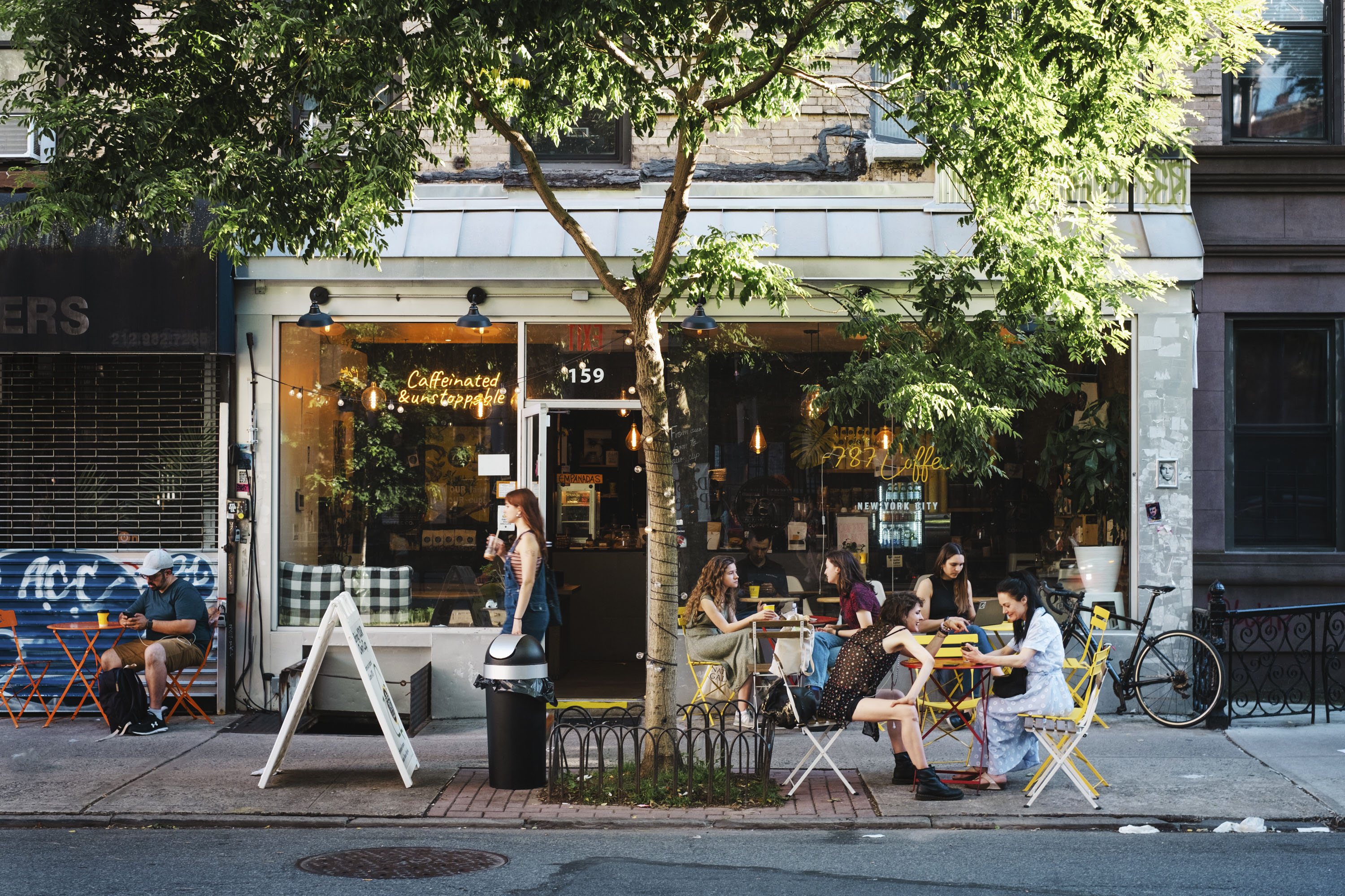 People relaxing at sidewalk cafe tables outside a New York coffee shop on a tree-lined street