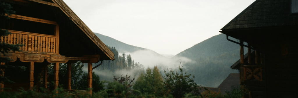 Wooden cabins frame misty forested mountains under a cloudy sky.