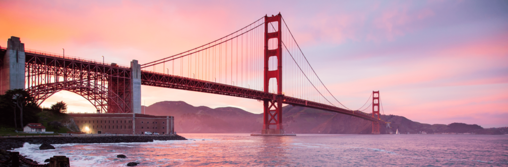 The Golden Gate Bridge at sunset, with a vibrant pink and orange sky over the bay. The iconic red suspension bridge spans the water, with a historic fort visible on the left side.