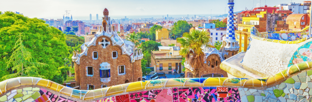 A view from Park Güell in Barcelona. The foreground shows a colorful, mosaic bench with two of the park