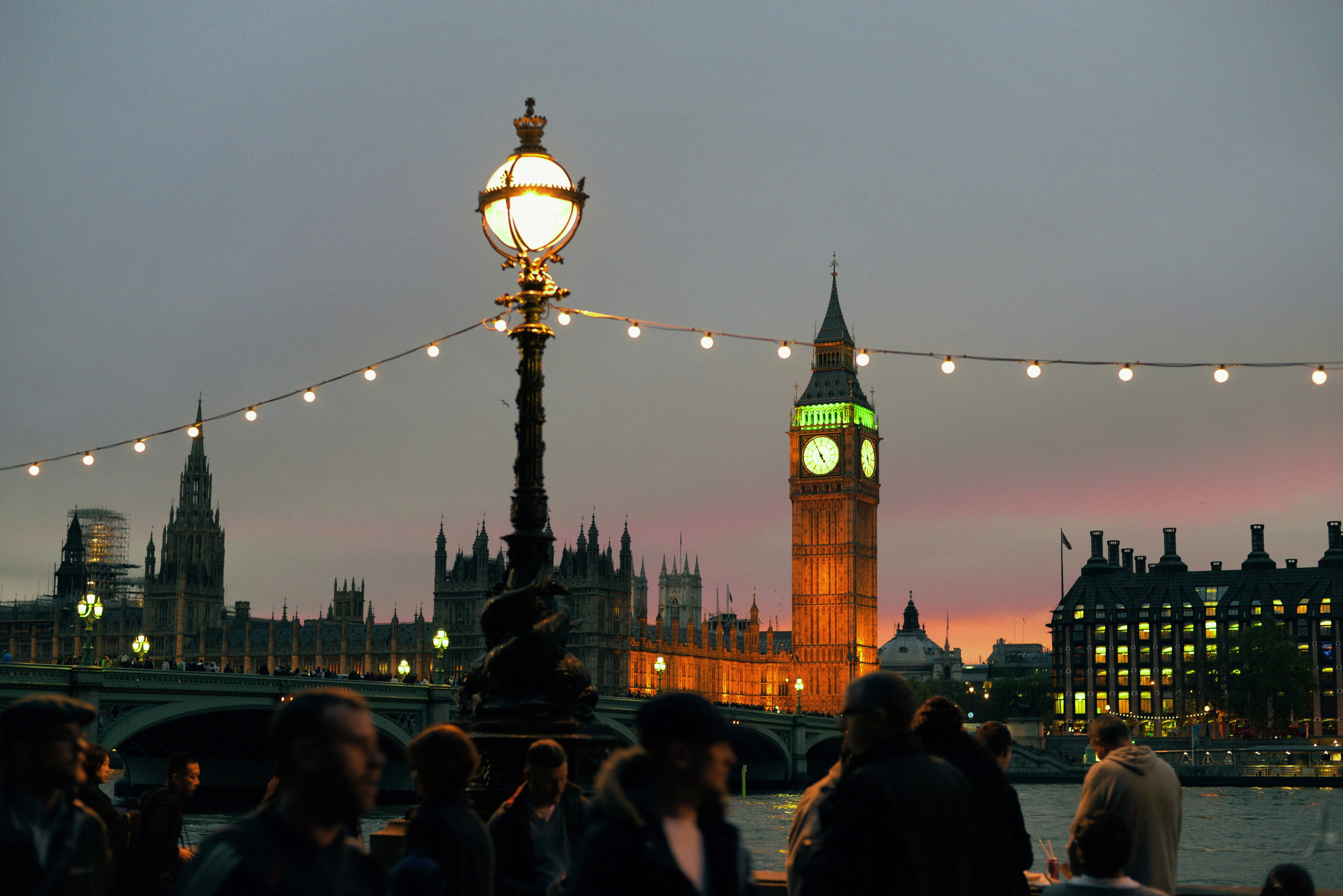 Big Ben and the Houses of Parliament lit at dusk, viewed across the Thames past a lamppost strung with festoon lights, London