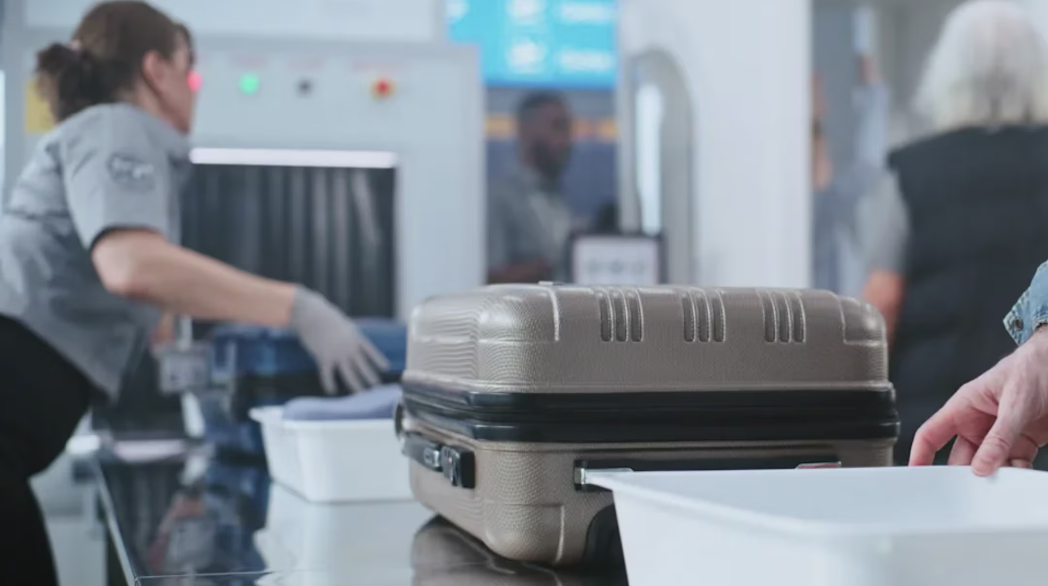 Airport security officer screening a carry-on suitcase and bins at a checkpoint conveyor
