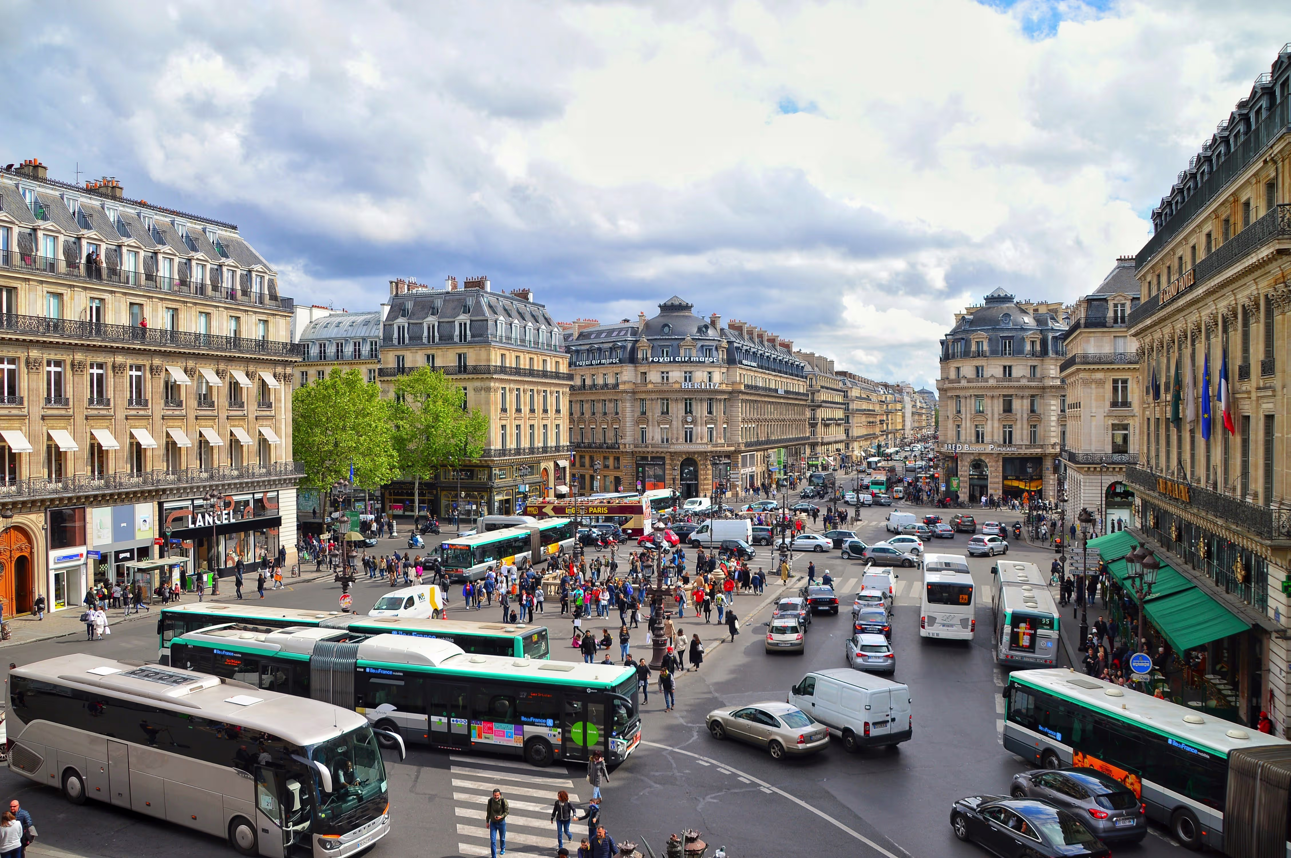 Busy Paris square near the Opera with buses, traffic and pedestrians among Haussmann buildings under a cloudy sky