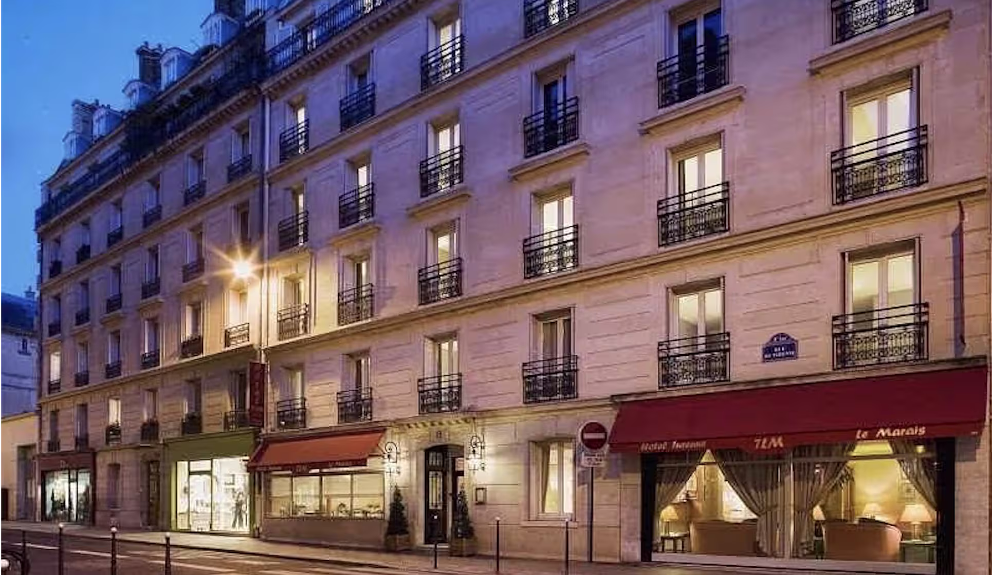 Classic Parisian Haussmann building lit at dusk with wrought-iron balconies and a hotel storefront on the ground floor